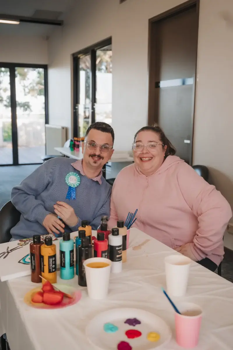 Participant and support worker smiling together at a table with paints